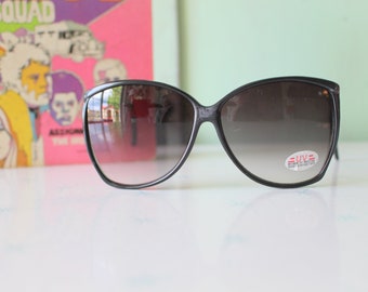 Puede incluir: Gafas de sol negras de ojo de gato con lentes de degradado marrón. Las gafas tienen una etiqueta "UV" en la lente derecha.