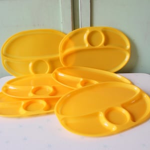 Op de afbeelding: Een verzameling felgele verdeelde borden. Elk bord heeft twee compartimenten en een cirkelvormige inkeping voor een kopje of kom. De borden zijn gemaakt van glanzend plastic en zijn gerangschikt op een wit oppervlak.