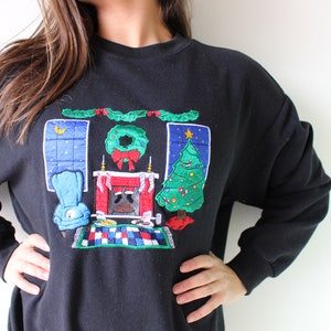 May include: Black sweatshirt with an embroidered Christmas scene featuring a fireplace, a Christmas tree, a wreath, and a chair. The scene is surrounded by a garland of holly.