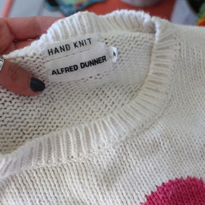 May include: White hand-knitted sweater with a pink and blue design. The sweater has a label that reads "HAND KNIT" and "ALFRED DUNNER".