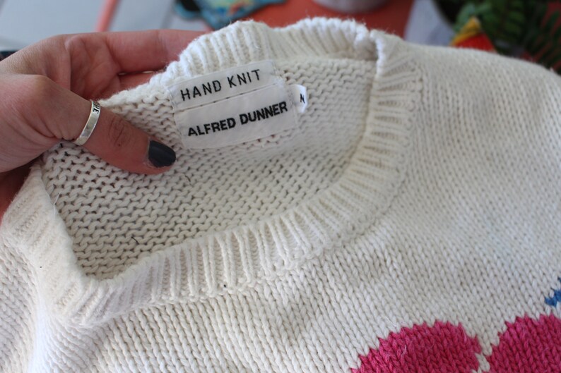 May include: White hand-knitted sweater with a pink and blue design. The sweater has a label that reads "HAND KNIT" and "ALFRED DUNNER".