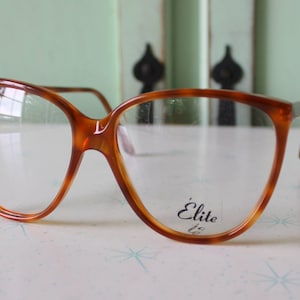Puede incluir: Gafas vintage de carey marrón con lentes transparentes. Las gafas tienen una montura grande y angular con la palabra "Élite" impresa en la lente. Las monturas son de color marrón cálido con un patrón de carey.