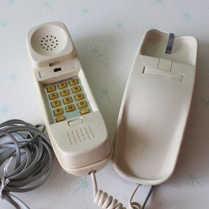 May include: A white rotary phone with a beige keypad and a coiled cord. The phone has a separate handset.