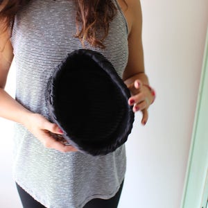 Puede incluir: Una cesta redonda de tela negra con una superficie texturizada. La cesta est&aacute; sostenida por una persona que lleva una camiseta sin mangas a rayas. La cesta parece estar vac&iacute;a.