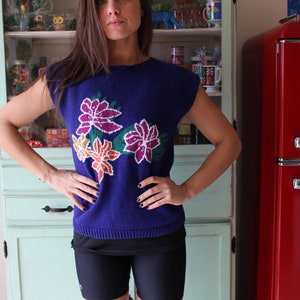 Op de afbeelding: Een vrouw draagt een blauwe trui met een bloemenpatroon. De trui heeft een ronde hals en korte mouwen. De bloemen zijn in tinten roze, oranje en geel.