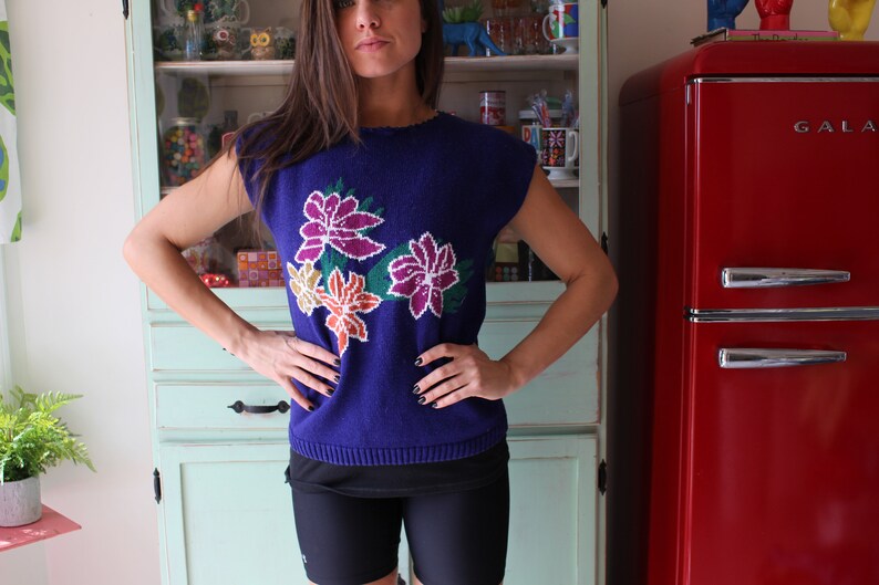 Op de afbeelding: Een vrouw draagt een blauwe trui met een bloemenpatroon. De trui heeft een ronde hals en korte mouwen. De bloemen zijn in tinten roze, oranje en geel.