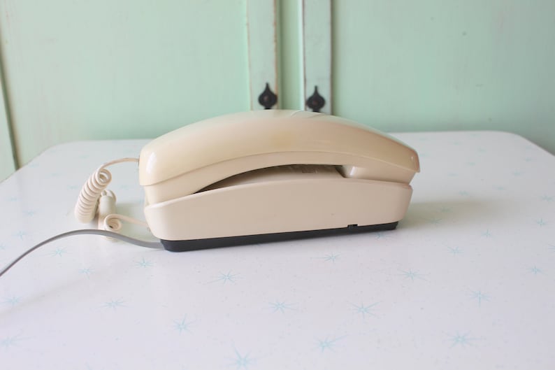 May include: A beige rotary phone with a coiled cord. The phone is on a white surface with a blue star pattern.