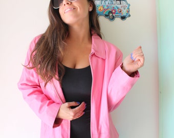 Puede incluir: Chaqueta rosa brillante con cuello y cierre de cremallera, sobre un top negro. La imagen también muestra una decoración de pared de una furgoneta con un símbolo de la paz.
