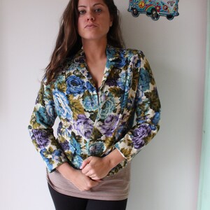 Puede incluir: Una mujer lleva una chaqueta de flores con flores azules, verdes y moradas sobre un fondo crema. La chaqueta tiene cuello y mangas largas.