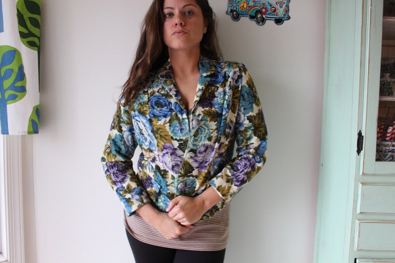 Puede incluir: Una mujer lleva una chaqueta de flores con flores azules, verdes y moradas sobre un fondo crema. La chaqueta tiene cuello y mangas largas.