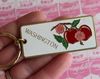 Puede incluir: Llavero rectangular con fondo blanco y borde dorado. El llavero presenta la palabra "WASHINGTON" en letras doradas, junto con un gráfico de manzanas rojas y flores rosas. Se adjunta un llavero dorado.