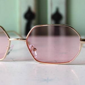 May include: Gold-framed sunglasses with a unique, geometric shape and pink-tinted lenses. The glasses have a delicate, wire-thin frame and are set against a soft, blurred background. The lenses are a soft pink color.