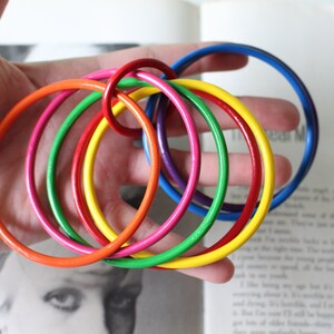 May include: A set of five interlocking bangle bracelets in vibrant solid colors including orange, green, yellow, purple, and blue. The circular bangles have a smooth, glossy finish and are linked together to form a single connected chain structure.