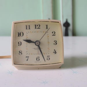 Peut inclure: Un réveil vintage blanc avec des chiffres et des aiguilles noirs. L'horloge a un cordon et est réglée sur 7h45.