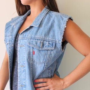 May include: A light blue denim vest with a red tab on the pocket. The vest has a collar and is sleeveless.