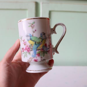 May include: A white ceramic mug with a gold rim and a footed base. The mug features a floral design with two blue, yellow, and green birds.
