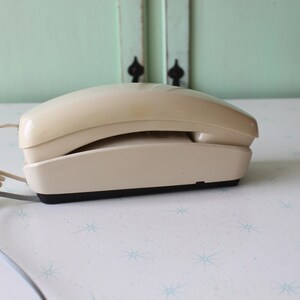 May include: A beige rotary phone with a coiled cord. The phone is on a white surface with a star pattern.