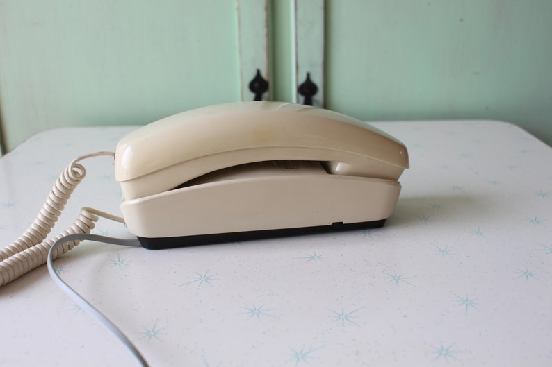 May include: A beige rotary phone with a coiled cord. The phone is on a white surface with a star pattern.