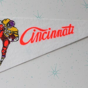 Pode incluir: Um flâmula de feltro branco com uma borda vermelha e a palavra "Cincinnati" em letras cursivas vermelhas. Uma ilustração de desenho animado de um jogador de futebol americano vestindo um uniforme amarelo e roxo está no lado esquerdo da flâmula.