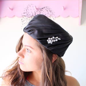 Puede incluir: Un sombrero tipo turbante de sat&eacute;n negro con una broche de pedrer&iacute;a. El sombrero presenta un dise&ntilde;o plisado y un delicado detalle de redecilla negra. Este tocado de inspiraci&oacute;n vintage a&ntilde;ade un toque de elegancia.