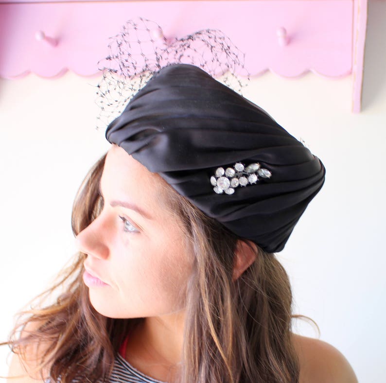 Puede incluir: Un sombrero tipo turbante de sat&eacute;n negro con una broche de pedrer&iacute;a. El sombrero presenta un dise&ntilde;o plisado y un delicado detalle de redecilla negra. Este tocado de inspiraci&oacute;n vintage a&ntilde;ade un toque de elegancia.