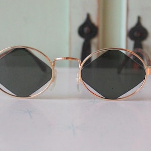 May include: Gold-rimmed sunglasses with dark, diamond-shaped lenses. The round frames have a thin gold metal and a bridge. The sunglasses are set against a light green background.