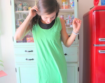 May include: A woman wearing a bright green sleeveless dress with a black undershirt. She is standing in front of a white cabinet with glass doors and a red refrigerator.