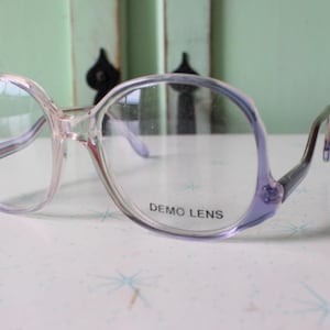 May include: Clear eyeglasses with a light purple frame. The glasses have a large, rounded shape with a subtle cat-eye design. The lenses are marked with the text "DEMO LENS".