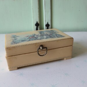 May include: A vintage cream-colored trinket box with a gold trim and a metal handle. The lid features a colorful illustration of a scene with a building and people.