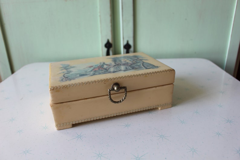 May include: A vintage cream-colored trinket box with a gold trim and a metal handle. The lid features a colorful illustration of a scene with a building and people.