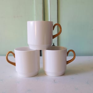 Könnte beinhalten: Drei weiße Keramikbecher mit goldenen Griffen. Die Becher sind übereinander gestapelt.