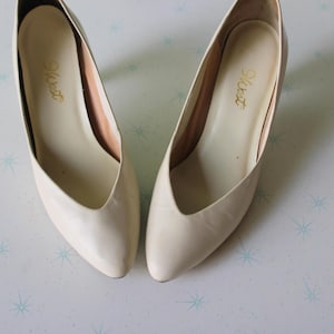 Könnte beinhalten: Ein Paar cremefarbene Pumps mit spitzer Zehenpartie. Die Schuhe haben einen kleinen Absatz und einen V-förmigen Vamp. Die Innenseite jedes Schuhs ist hellbraun, mit dem Wort "West" in goldener Schrift. Wahrscheinlich Vintage.