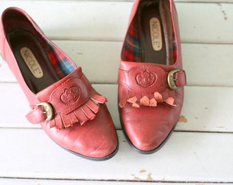 May include: A pair of red leather shoes with a fringe detail and a buckle fastening. The shoes have a plaid lining and the brand name "NICOLE" is printed on the inside.