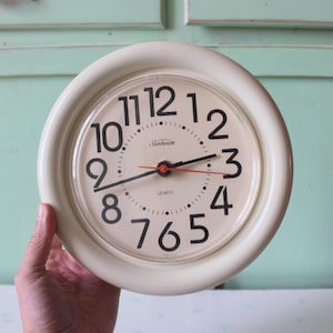 May include: A round wall clock with a cream-colored frame and a matching cream face. The clock features large black Arabic numerals, black hour and minute hands, and a thin red second hand. The word Sunbeam is printed in black above the center.
