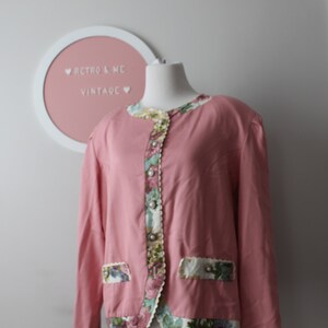 May include: A pink long-sleeved blouse with a floral print trim around the neckline, cuffs, and hem. The blouse has a button closure and a pocket on the left side.
