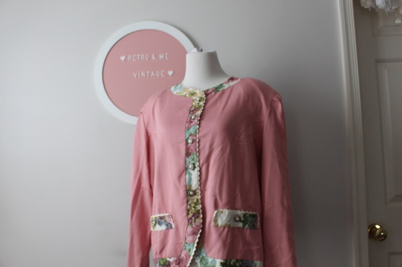 May include: A pink long-sleeved blouse with a floral print trim around the neckline, cuffs, and hem. The blouse has a button closure and a pocket on the left side.