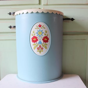 Könnte beinhalten: Ein hellblaues Metall-Abfalleimer mit einem wellenförmigen weißen Rand. Ein ovales Blumendesign mit roten, gelben und blauen Blumen ist mittig auf der Vorderseite. Der Abfalleimer ist zylindrisch und hat einen abgerundeten Boden.