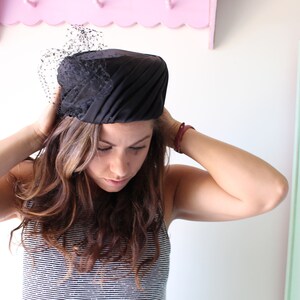 Puede incluir: Un sombrero negro con velo. El sombrero presenta un dise&ntilde;o estructurado y plisado, con un peque&ntilde;o y delicado velo en la parte delantera. Este sombrero de estilo vintage es adecuado para ocasiones especiales.