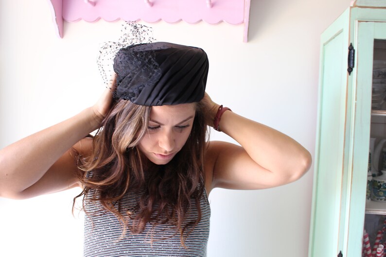 Puede incluir: Un sombrero negro con velo. El sombrero presenta un dise&ntilde;o estructurado y plisado, con un peque&ntilde;o y delicado velo en la parte delantera. Este sombrero de estilo vintage es adecuado para ocasiones especiales.