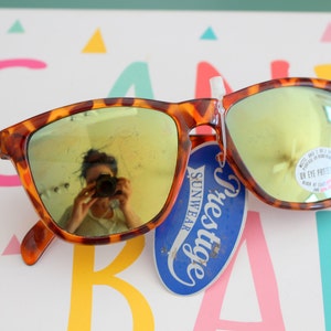 Może przedstawiać: Okulary przeciwsłoneczne w kolorze żółwim z lustrzanymi złotymi szkłami i niebieską metką z napisem "Prestige Sunwear".