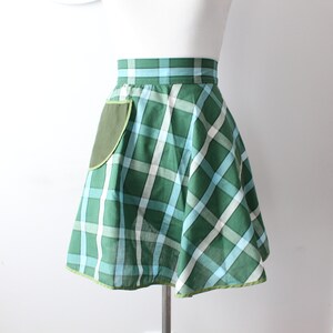 Pode incluir: Um avental xadrez verde e azul com um bolso verde. O avental tem uma saia franzida e uma bainha verde.