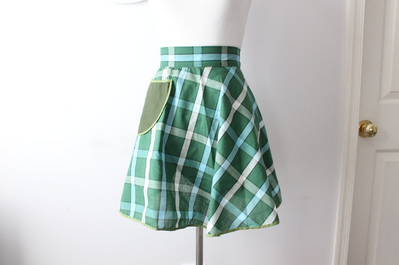 Pode incluir: Um avental xadrez verde e azul com um bolso verde. O avental tem uma saia franzida e uma bainha verde.