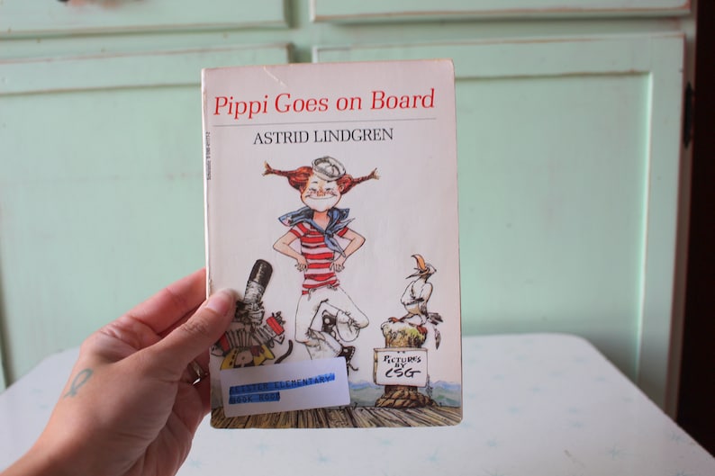 May include: A vintage children's book titled "Pippi Goes on Board" by Astrid Lindgren. The cover features a colorful illustration of Pippi Longstocking, a fictional character with red pigtails, wearing a sailor outfit and standing on a boat deck. The book has a white background with red and black text.