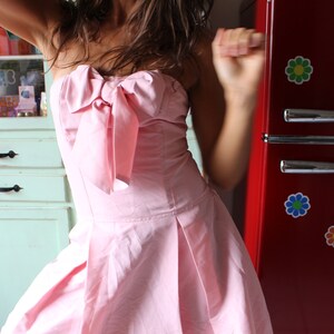 May include: A woman in a pink strapless dress with a large bow at the bust is dancing in front of a red refrigerator with flower decals.