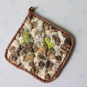 Puede incluir: Un agarrador de olla acolchado marrón con fondo blanco y un patrón de ranas verdes y champiñones marrones.