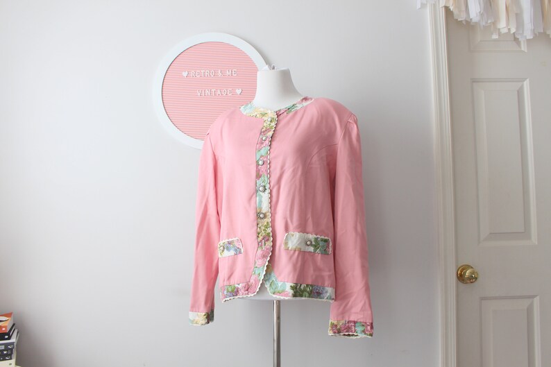 May include: A pink vintage jacket with floral trim and button details. The jacket has a scalloped hem and long sleeves.