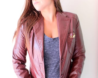 May include: A burgundy leather blazer with a gold pin on the left lapel. The blazer has a single button closure and two pockets.