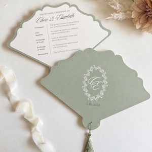 May include: A sage green fan-shaped wedding program with a scalloped edge. The front features a white floral wreath monogram with the letters CE and the date 12.05.2028. The interior lists the wedding ceremony order for Clare and Elizabeth.