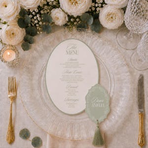May include: An oval menu card with white paper and sage green border, featuring the text The Menu, Starters, Main Course, Dessert, and Beverages. A sage green name tag with the text Dear Amelia and a matching tassel rests on a clear glass plate.