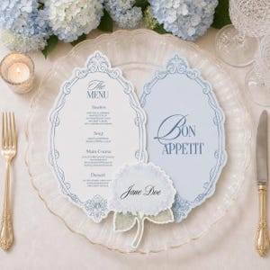 May include: A white menu card with blue ornate borders and text, a light blue card with the text Bon Appetit, and a small hydrangea-shaped place card with the name Jane Doe, all arranged on a clear glass plate with a gold fork and knife.